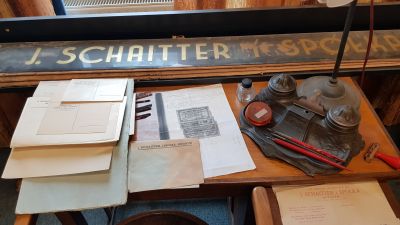 Prywatny sekretarzyk z kancelarii Michała Franciszka Gottmana, jego przybornik do pisania, lampka, bony towarowe, firmowe koperty. U g&oacute;ry widoczny napis stanowiący element aranżacji witryny sklepowej. Zdjęcie udostępnione przez Pana Grzegorza Dąbrowskiego